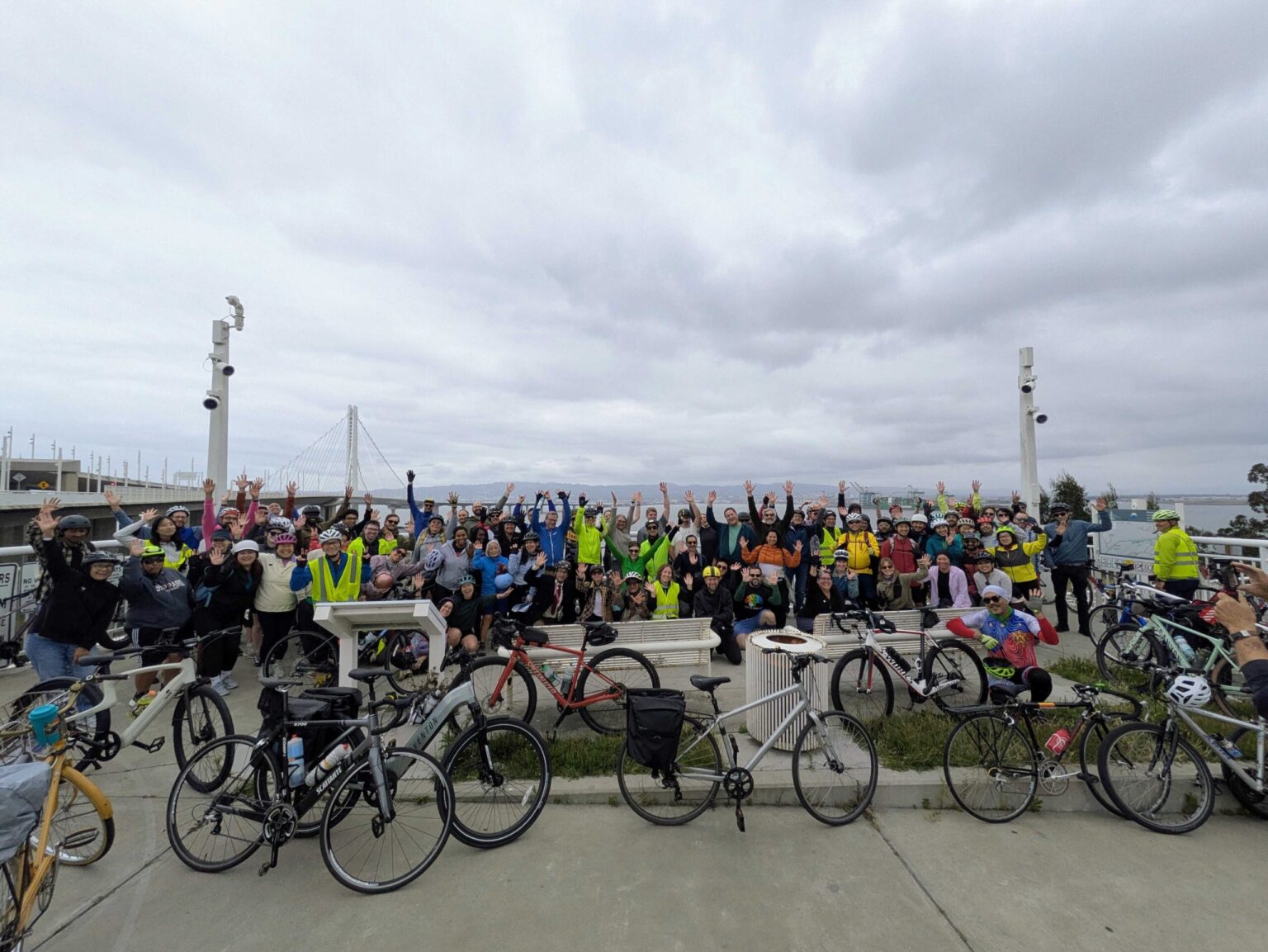 A large, diverse group of dozens of people. Many people in the group have their hands raised in celebration. In the foreground, several bicycles of various types—including road, hybrid, and mountain bikes—are parked or leaning against low concrete walls. In the background, the Bay Bridge spans across the water toward a distant City of Oakland skyline.