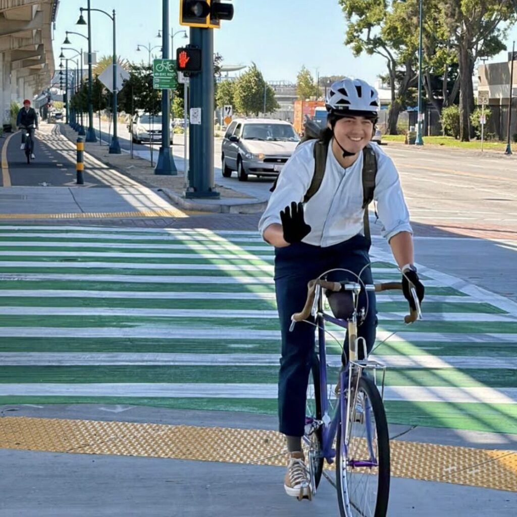 photo of a person riding a bike while smiling and waving, on the East Bay Greenway north of Coliseum BART in Oakland