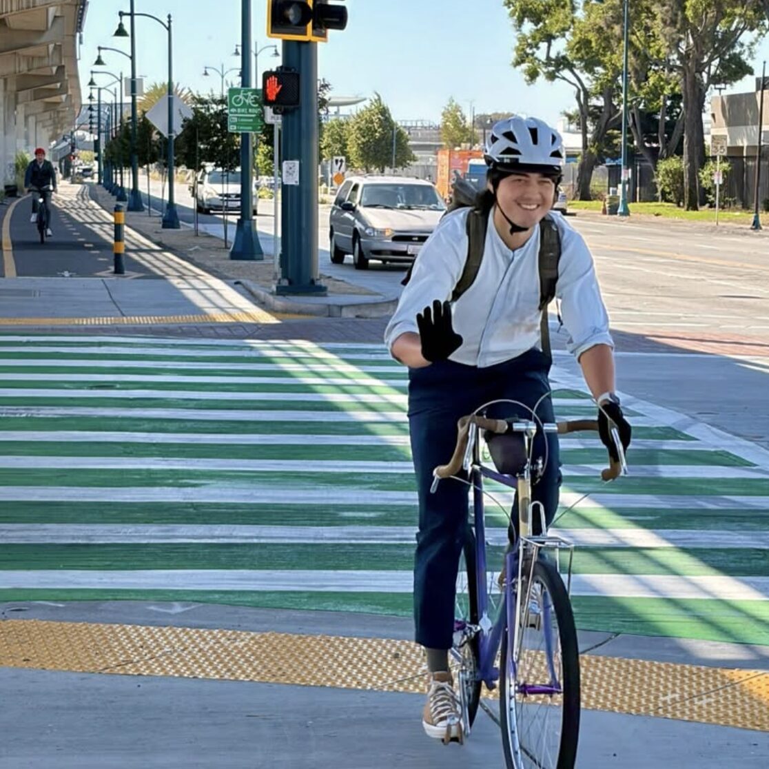 photo of a person riding a bike while smiling and waving, on the East Bay Greenway north of Coliseum BART in Oakland