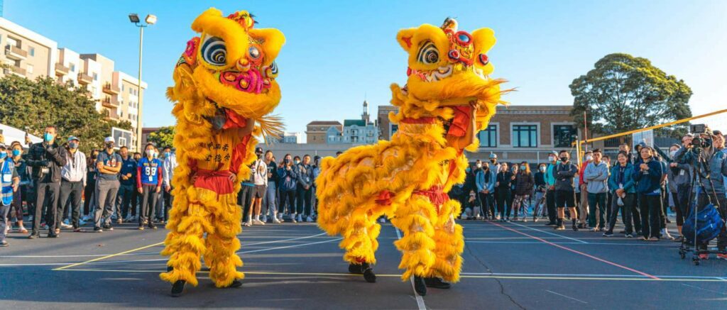 Photo of Chinese dragon dancers performing in front of a large crowd, outside on a basketball court