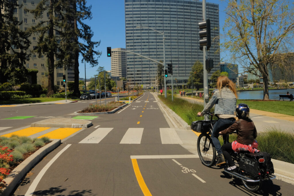 Photo of a parent on a cargo bike with a helmeted child riding on the back, in the two-way protected cycletrack on Lakeside Ave in Oakland