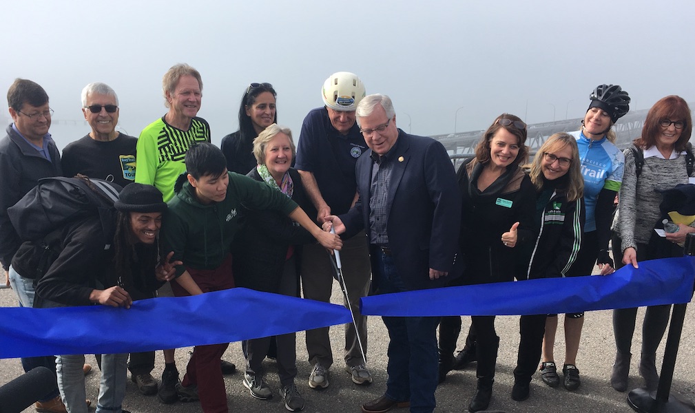 photo of a group of people cutting a large ribbon with a pair of large scissors, with the Richmond-San Rafael Bridge in the background