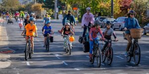 A group of mostly kids with some adults on a group bike ride on the street wearing Halloween costumes