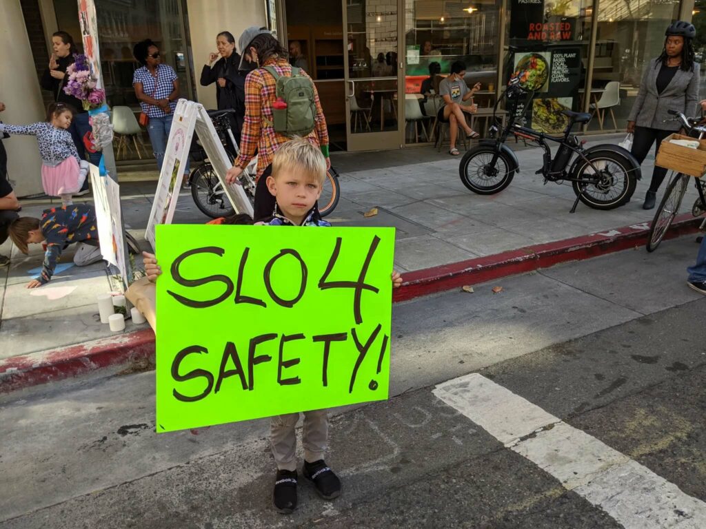 Photo of a child holding a sign "slo 4 safety" at a vigil for a bike rider killed in a crash on 14th St in Oakland