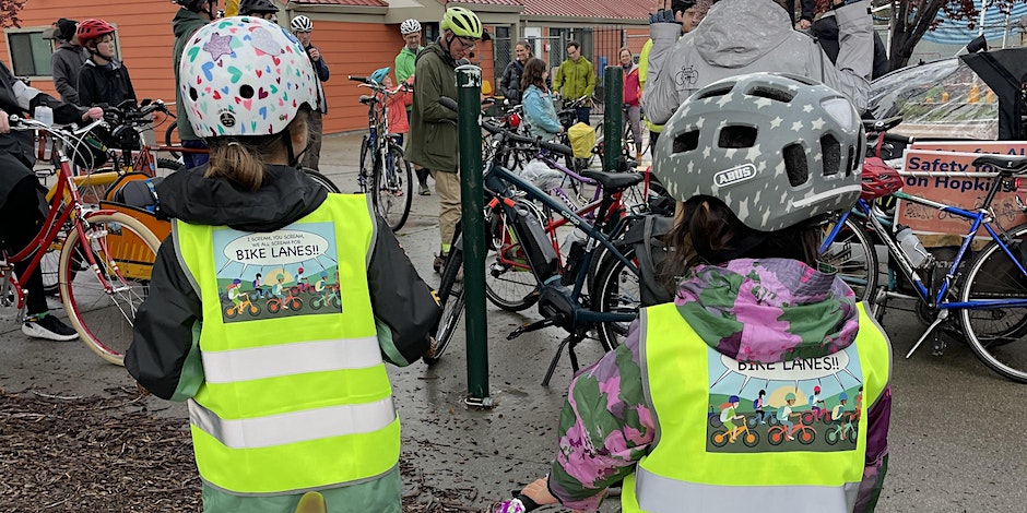 Photo from the back of a group of bike riders, with two children wearing helmets and high viz vests printed with a cartoon graphic of a group of bike riders and a word bubble "bike lanes!"