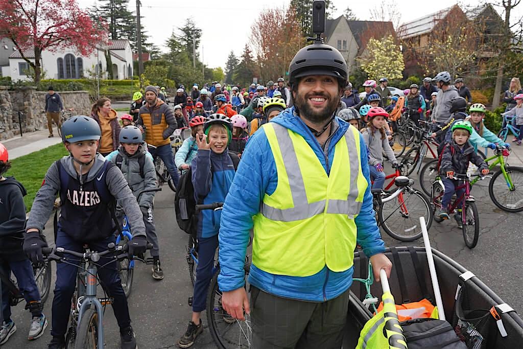 Coach Balto stands in front of a large crowd of young students wearing helmets and riding bicycles.