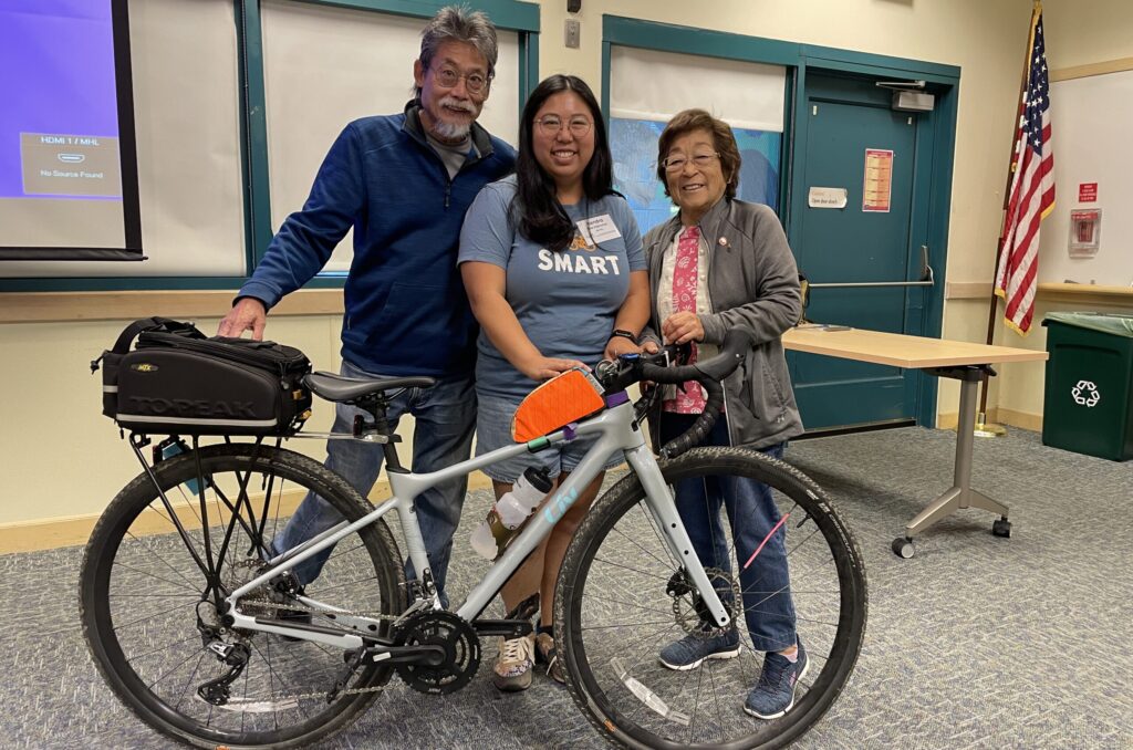 Kendra poses with her dad and grandma, all standing behind her bike as she demonstrates bike tips as a class instructor.