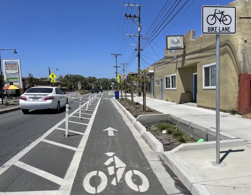 photo of a curbside bicycle lane on Rumrill Blvd in the City of San Pablo CA, with a painted buffer space to the left with flex posts separating the bikeway from a car in the travel lane, and a concrete rain garden and young trees in a landscaping area to the right