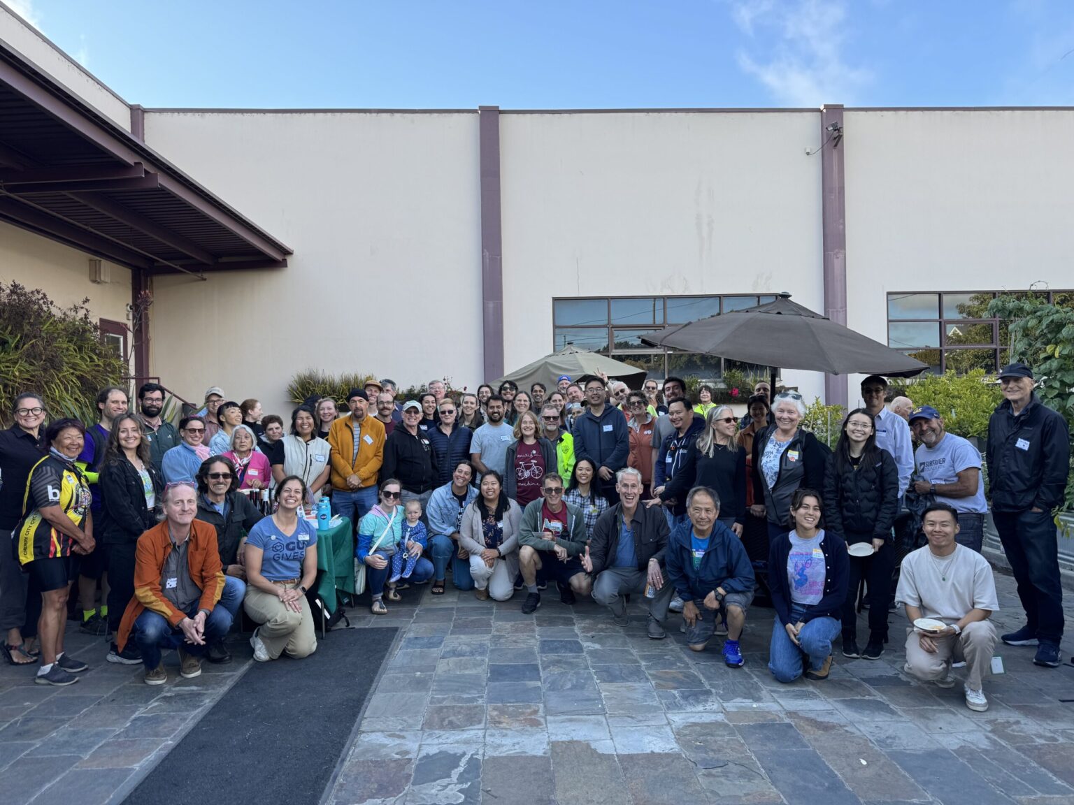 Group photo of about 60 people smiling in the GU Backyard, all dressed in relaxed summer outfits