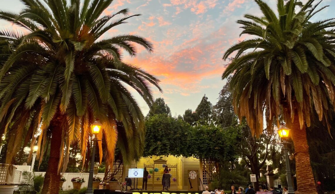 An evening shot of an outdoor event with a stage and seating. The sky is a gradient of orange, pink, and blue at sunset. Two large palm trees with fronds framing the top of the photo are lit by warm lamps. People are sitting at tables in the grassy area to the right, and two people are on a stage with a screen and a sign that says **BIKETOPIA**. There are a few trees in the background, and the overall atmosphere is peaceful.