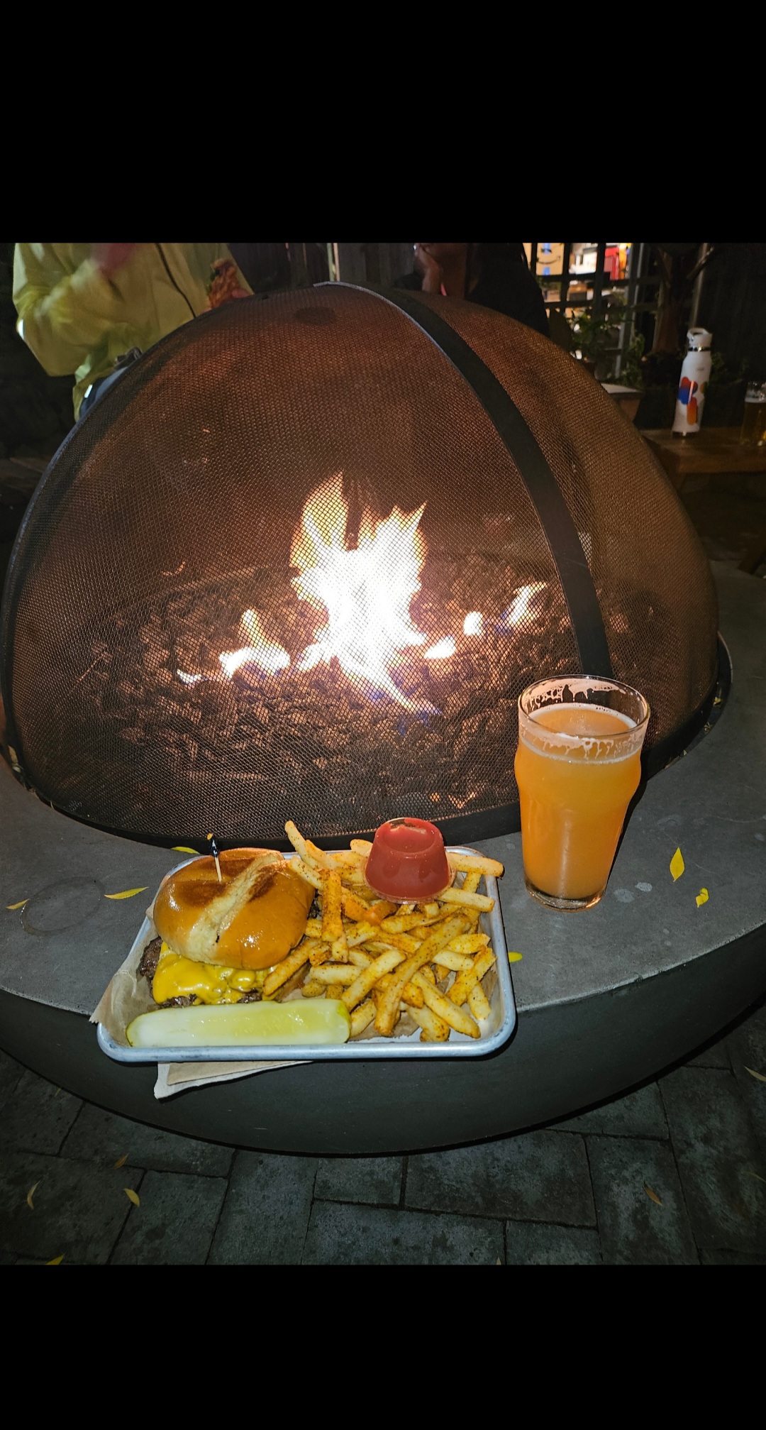 A metal tray with a cheeseburger, french fries, ketchup, and a pickle spear sits on the edge of a large, circular fire pit. To the right of the tray is a glass of hazy, light-colored beer. Inside the fire pit, which is covered by a mesh dome, bright orange flames are visible. The scene is outdoors at night, lit by the fire.