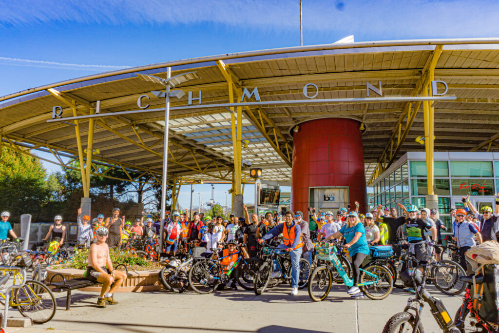 A large group of cyclists poses for a photo under the covered entrance of the Richmond BART station.