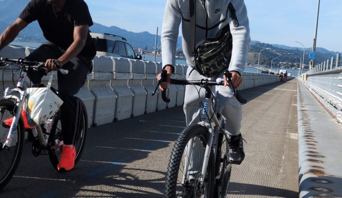 Two people are cycling side-by-side across a bridge on a sunny day