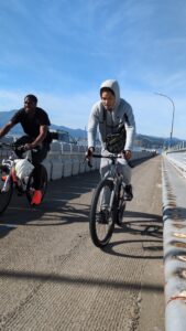 Two people are cycling side-by-side across a bridge on a sunny day