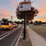 Photo of a painted buffer bike lane on a street next to cars - A sign mounted above the street reads "BART PEDESTRIAN and BICYCLE CONNECTIVITY ACTIVE TRANSPORTATION IMPROVEMENTS COMPLETION DATE: FALL 2024 YOUR TAX DOLLARS AT WORK BE WORK ZONE ALERT"