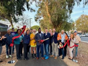 photo of Pittburg staff, city and regional electeds, and others standing on a paved and landscaped trail, cutting a large ribbon with a large pair of scissors