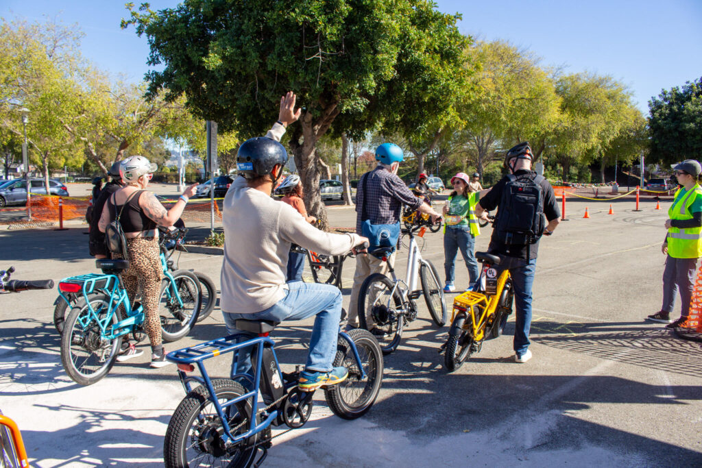 A group of people wearing helmets are gathered on a paved lot with several e-bikes and conventional bicycles, possibly at a demo or riding area. In the center foreground, a man in a light sweater and blue jeans, sitting on a blue cargo bike, has his right arm raised in the air. To his left, a woman wearing a leopard-print tank top stands next to a turquoise e-bike. Other people are visible talking and standing by their bikes, including one person next to a bright yellow e-bike with a cargo rack. The area is lined with trees and orange cones, with parked cars visible in the far background.