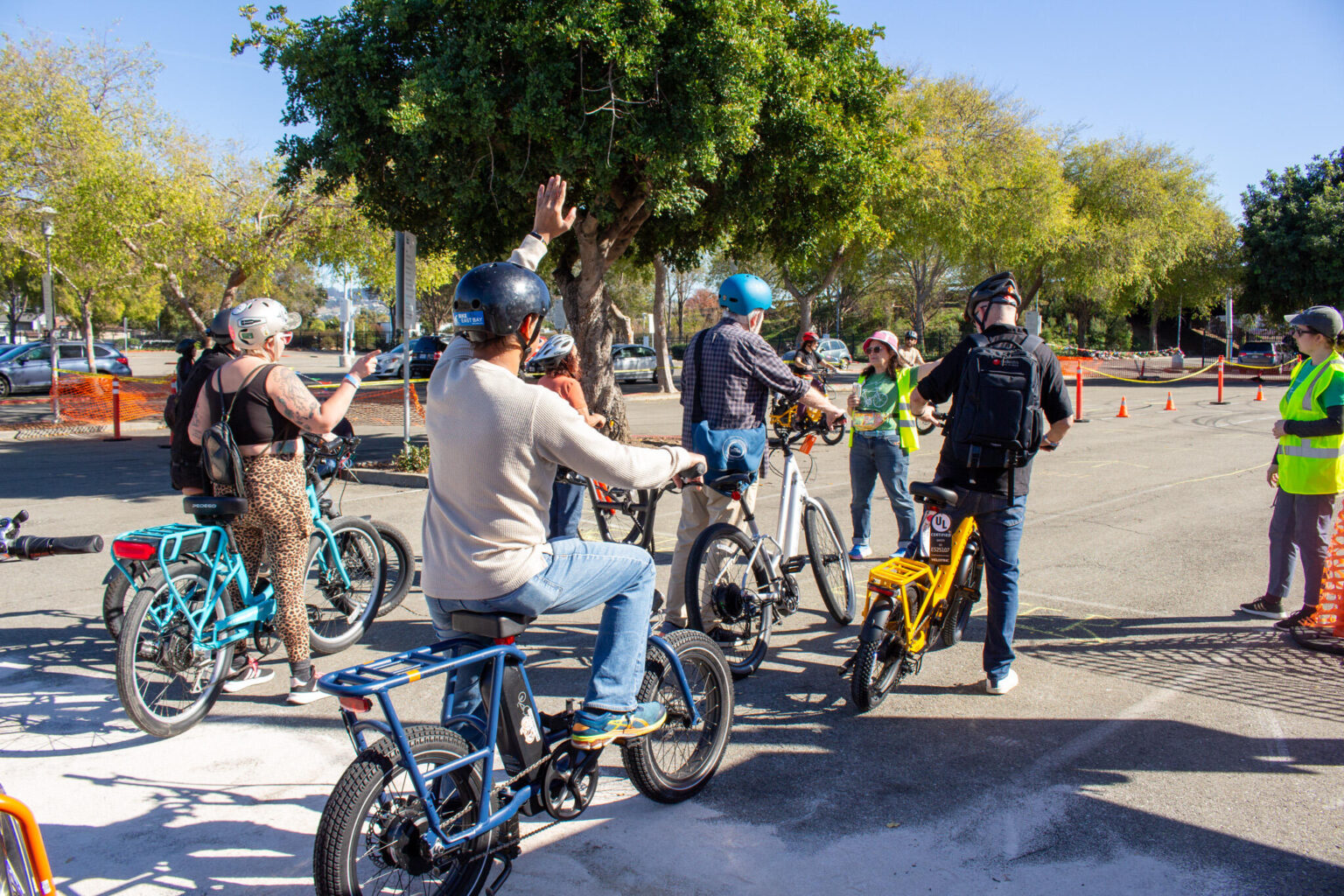 A group of people wearing helmets are gathered on a paved lot with several e-bikes and conventional bicycles, possibly at a demo or riding area. In the center foreground, a man in a light sweater and blue jeans, sitting on a blue cargo bike, has his right arm raised in the air. To his left, a woman wearing a leopard-print tank top stands next to a turquoise e-bike. Other people are visible talking and standing by their bikes, including one person next to a bright yellow e-bike with a cargo rack. The area is lined with trees and orange cones, with parked cars visible in the far background.
