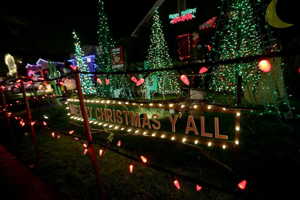 A nighttime photograph of a yard decorated with bright Christmas lights. In the foreground, a low fence is lined with red, cone-shaped lights. Behind the fence, a large wooden sign illuminated by white lights reads: "MERRY CHRISTMAS Y'ALL." The yard behind the sign features several small, cone-shaped Christmas trees densely wrapped in green lights. A darker house in the background also has some lights, including a red sign that says "Happy Holidays".