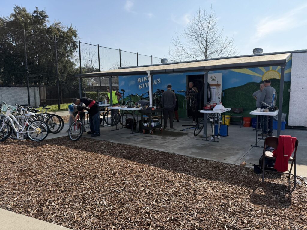 An outdoor scene at the Concord community bicycle repair workshop called "Bike Kitchen." Under a long metal awning attached to a blue shipping container muraled with hills and a rising sun, several people are actively working on bicycles. Three white folding tables serve as workstations, cluttered with tools and bike parts. A man in a high-visibility yellow vest stands near the center, while others lean over bikes in various states of repair. To the left, a row of bicycles is parked. The foreground consists of a large area covered in brown wood chips, and the weather appears bright and sunny under a clear blue sky. An empty folding chair with a red jacket draped over it sits in the lower right corner.