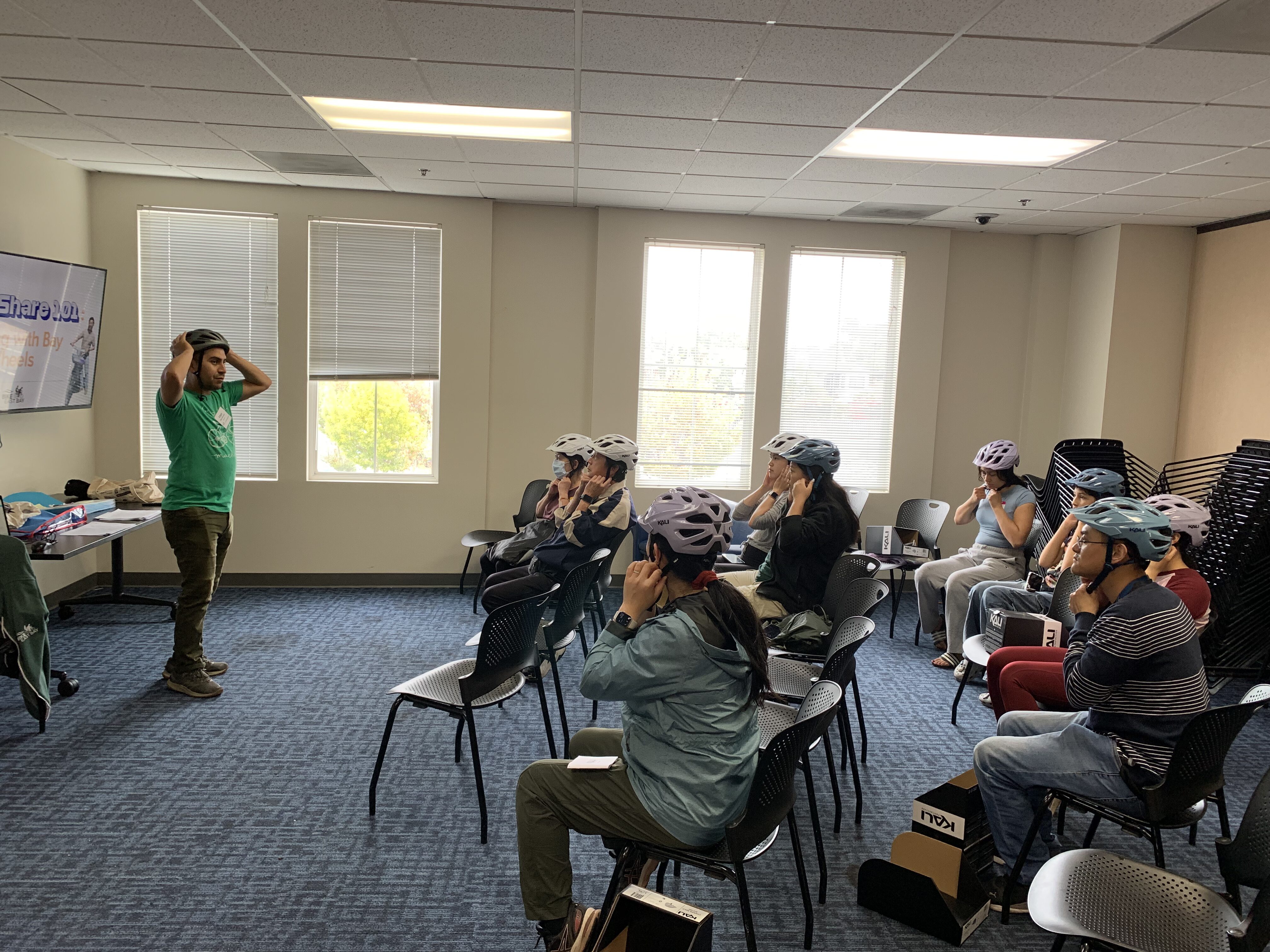 A group of people wearing helmets are seated in a classroom setting, as an instructor demonstrates how to properly fit a helmet.