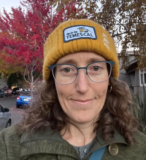 Shannon smiles in a selfie, wearing a yellow beanie with a pink tree in the background