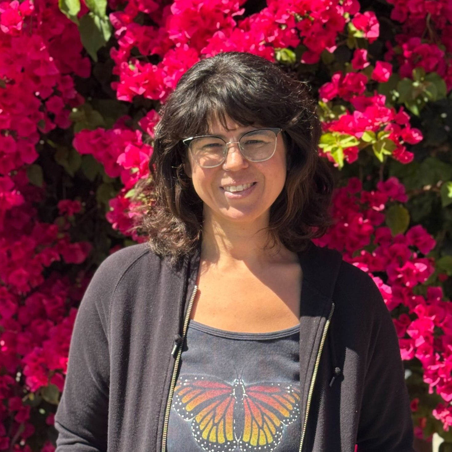 Leah is wearing a black shirt with a monarch, and a black hoodie, smiling in front of a blooming bougainvillea tree.
