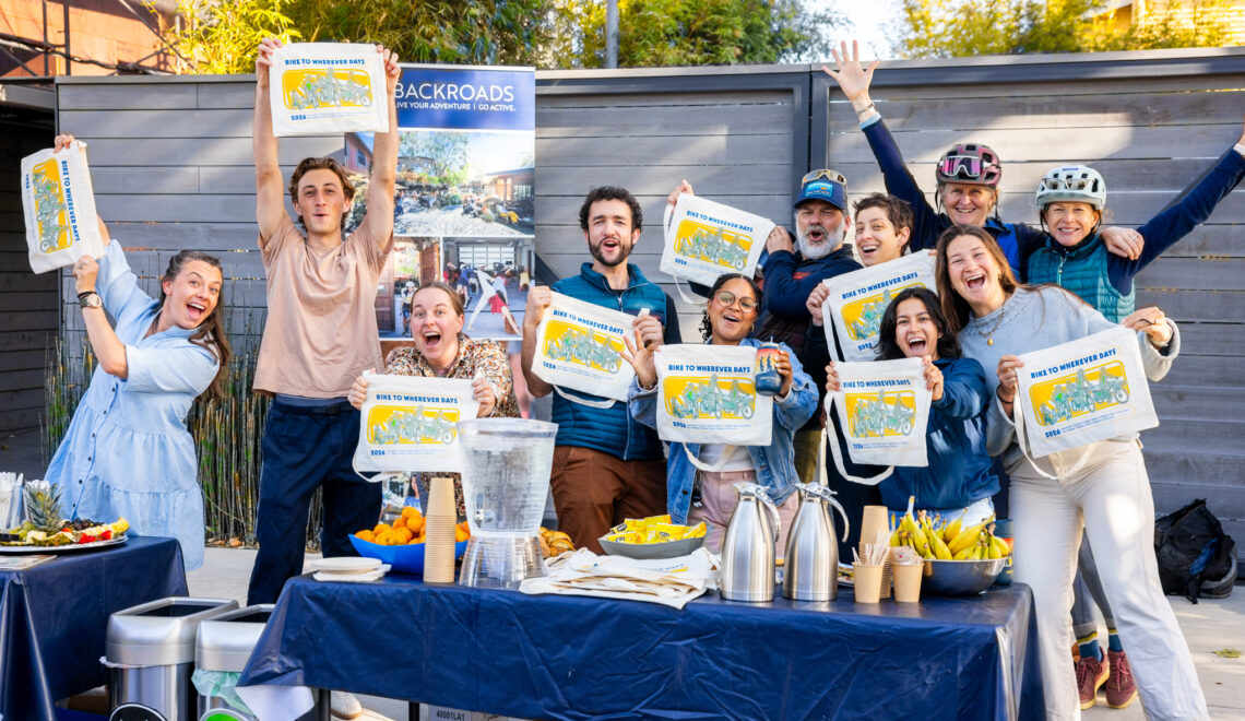 A group of smiling staff and volunteers at Backroads office in Berkeley smile and hold up BTWD bags.