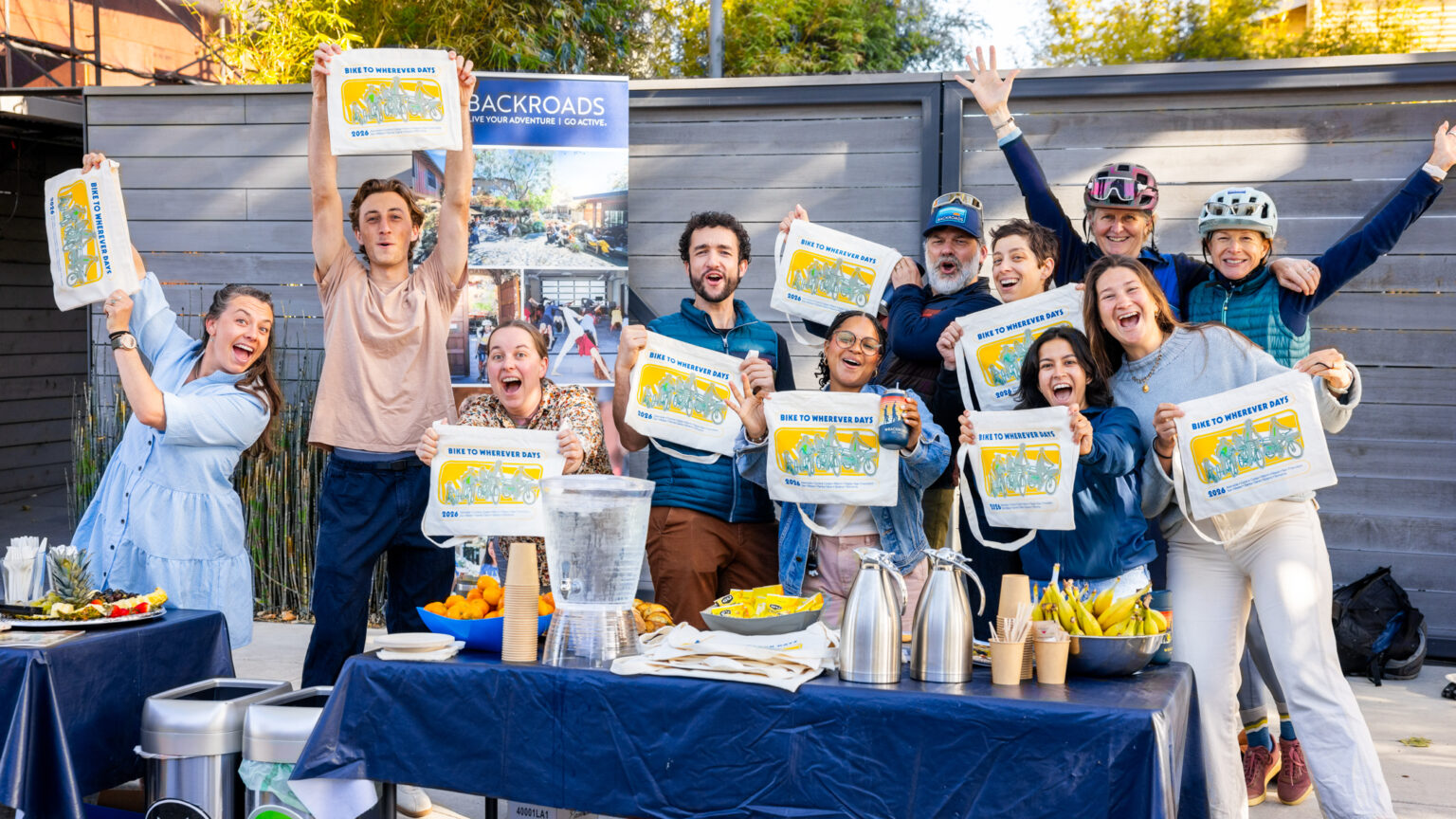A group of smiling staff and volunteers at Backroads office in Berkeley smile and hold up BTWD bags.