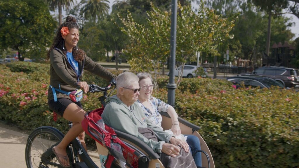 A side-profile shot of a woman with a flower in her hair pedaling a black electric trishaw along a sunlit park path. Two elderly passengers—a man in a grey hoodie and a woman in a floral blouse—sit side-by-side in the front carriage, looking ahead with smiles. The background features lush green bushes, palm trees, and a parked car under a bright, clear sky.