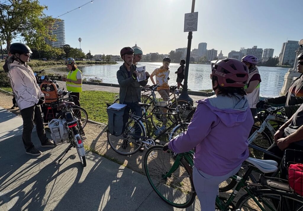 Photo of a group of people standing with their bikes and watching a person talk, standing next to Lake Merritt in Oakland