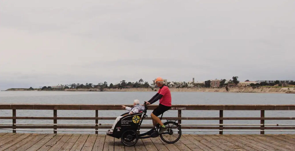 A man rides a specialized cargo tricycle with an elderly passenger on a wooden pier, with the ocean and a distant coastline under an overcast sky in the background.