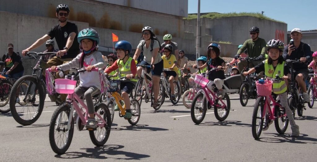 photo of a large group of youth bike riders with adults, riding past the Oakland Museum of California