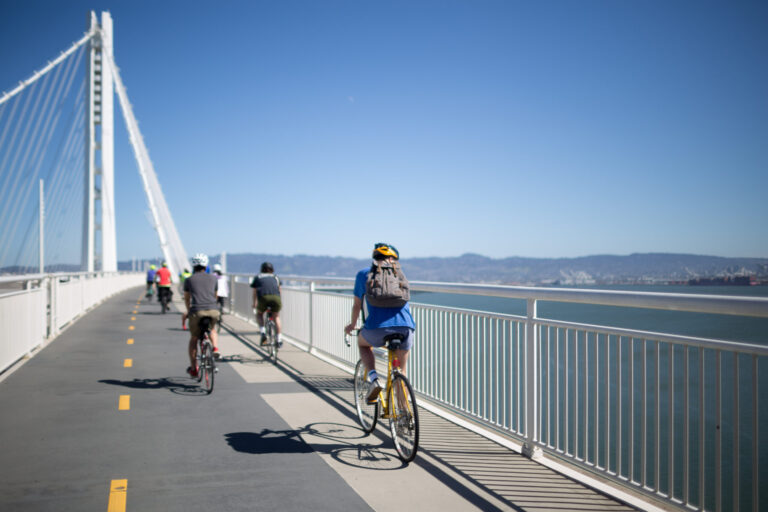 A group of people on bikes riding along the Alex Zuckerman bike and pedestrian path on the east span of the Bay Bridge under a clear blue sky, with water and distant hills in the background.